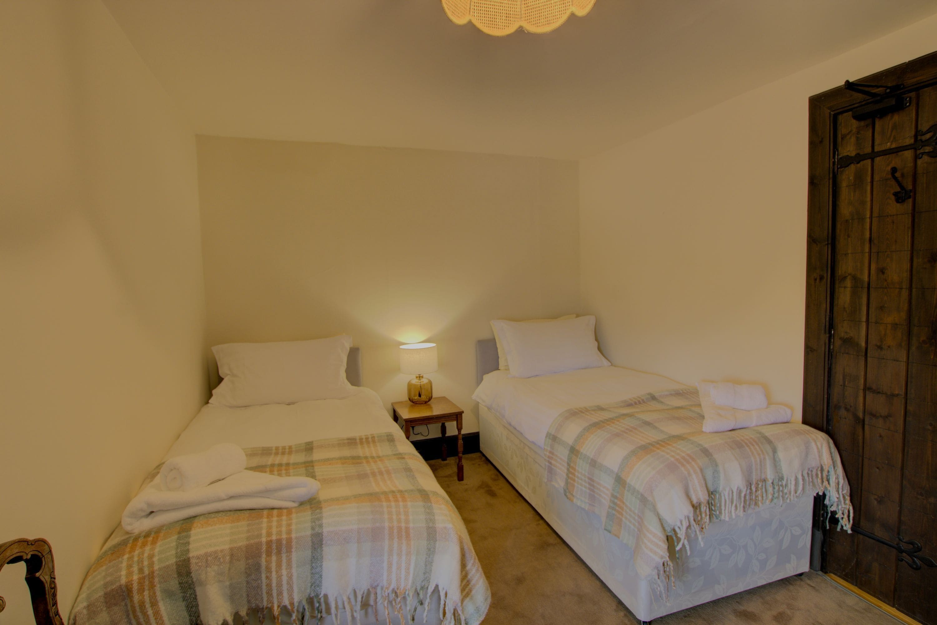A room with two single beds, each with a white pillow and a plaid blanket. There is a small bedside table with a lamp between the beds.