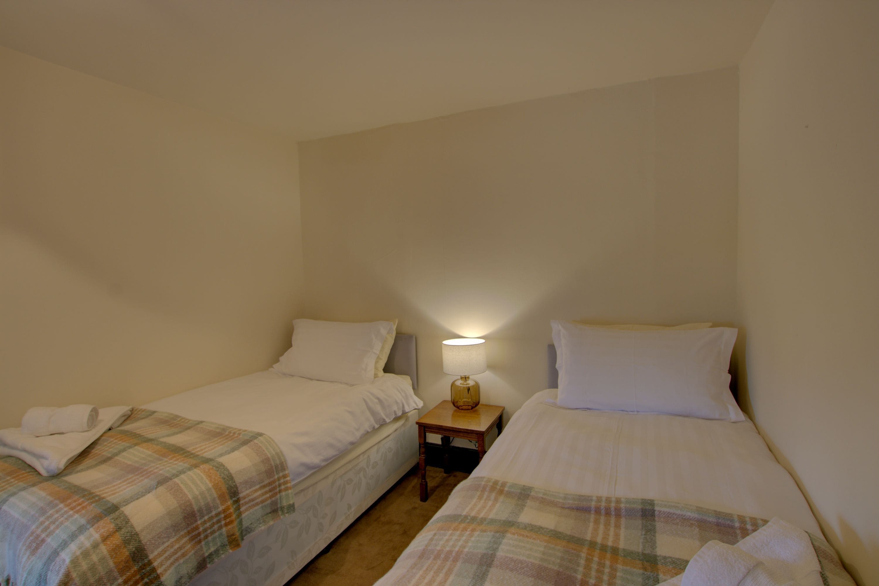 A small, single-room accommodation with two single beds, each with a plaid blanket and white pillows. There is a nightstand with a lamp between the beds.