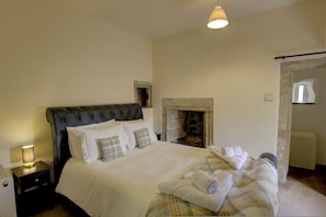 A bedroom with a large bed, a nightstand, a lamp, a fireplace, and a door leading to another room. - Little Tarrel Castle Portmahomack (Tain)