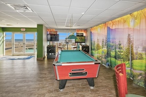 A pool table in a room with a view of the beach through the exit door, a television screen, and a mural of a landscape. - North Myrtle Beach Oceanview Condo (North Myrtle Beach)