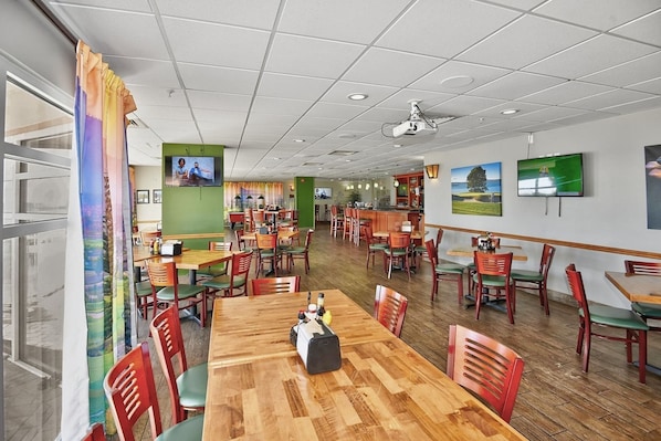 A spacious dining area with multiple tables and chairs, a television mounted on the wall, and a view of the outdoors through large windows. - North Myrtle Beach Oceanview Condo (North Myrtle Beach)