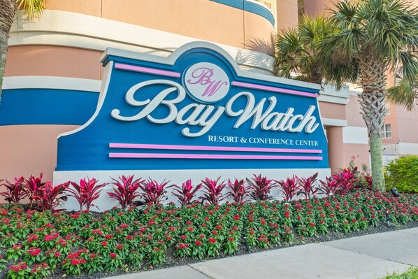 A sign for Bay Watch Resort & Conference Center with a decorative border and a row of red flowers in front. - North Myrtle Beach Oceanview Condo (North Myrtle Beach)