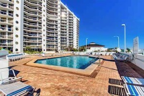 A hotel pool area with a rectangular pool, lounge chairs, and a high-rise building in the background. - Ocean View Resort Condo, Private Balcony, & Shared Hot Tub, Pool, Tennis (Miramar Beach)