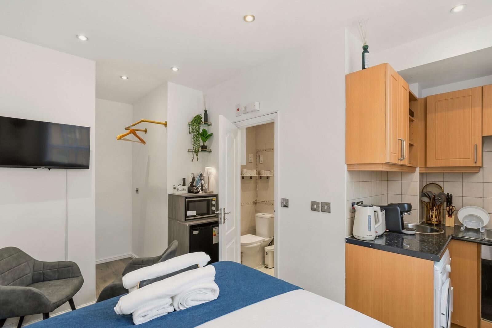 A compact hotel room with a bed, a desk, a chair, a kitchenette, and a bathroom.