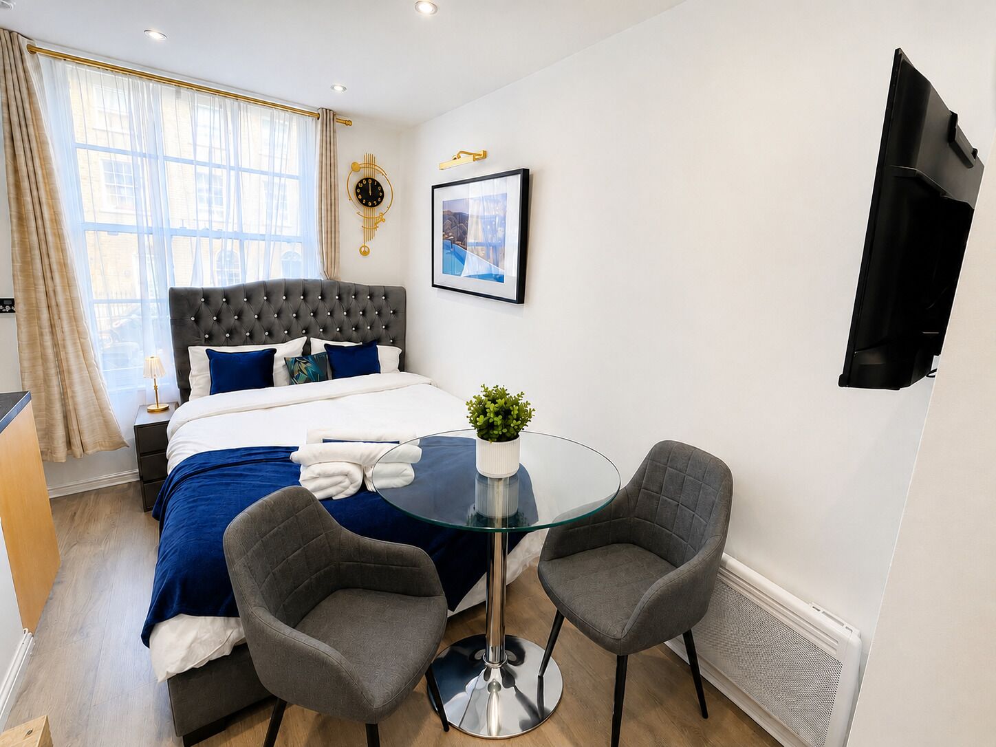 Habitación de hotel con cama, mesita de noche, lámpara, planta en maceta, dos sillas, una mesa redonda de vidrio y un televisor de pared.