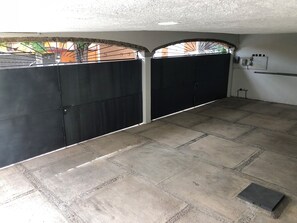 Un espace extérieur abrité, doté d’un sol carrelé, d’un portail en métal et d’une boîte électrique fixée au mur.