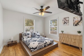 Une chambre à coucher avec un lit, un ventilateur de plafond, une commode en bois et des œuvres d’art accrochées au mur.