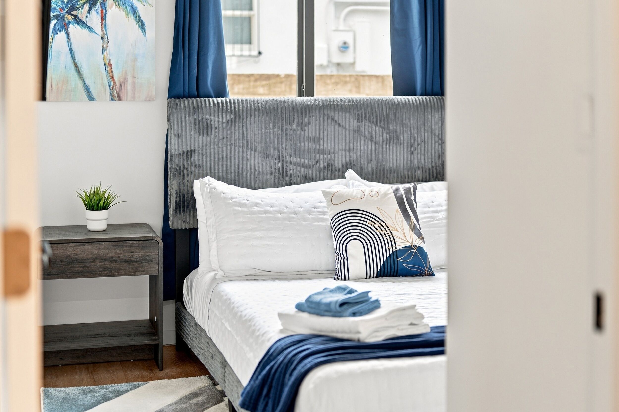 Een netjes opgemaakt bed met een gestreept kussen, een klein nachtkastje met een plant en een schilderij aan de muur.