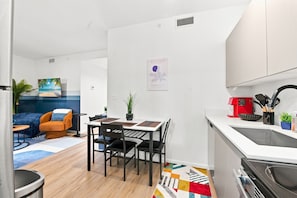 A modern kitchen with a dining area, a small living space with a sofa and a TV, and a colorful rug. - Cozy Fun and Vibrant Central Wynwood! King Bed! (Miami)