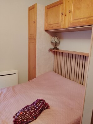 A single bed with a pink bedspread and a patterned blanket, a wooden cabinet above, and a radiator on the wall. - Ideal Vacation (Onnion)