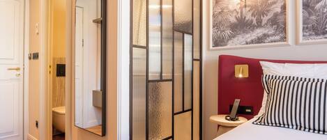 A hotel room with a bed, a bedside table with a lamp, a mirror, and a door leading to a bathroom.