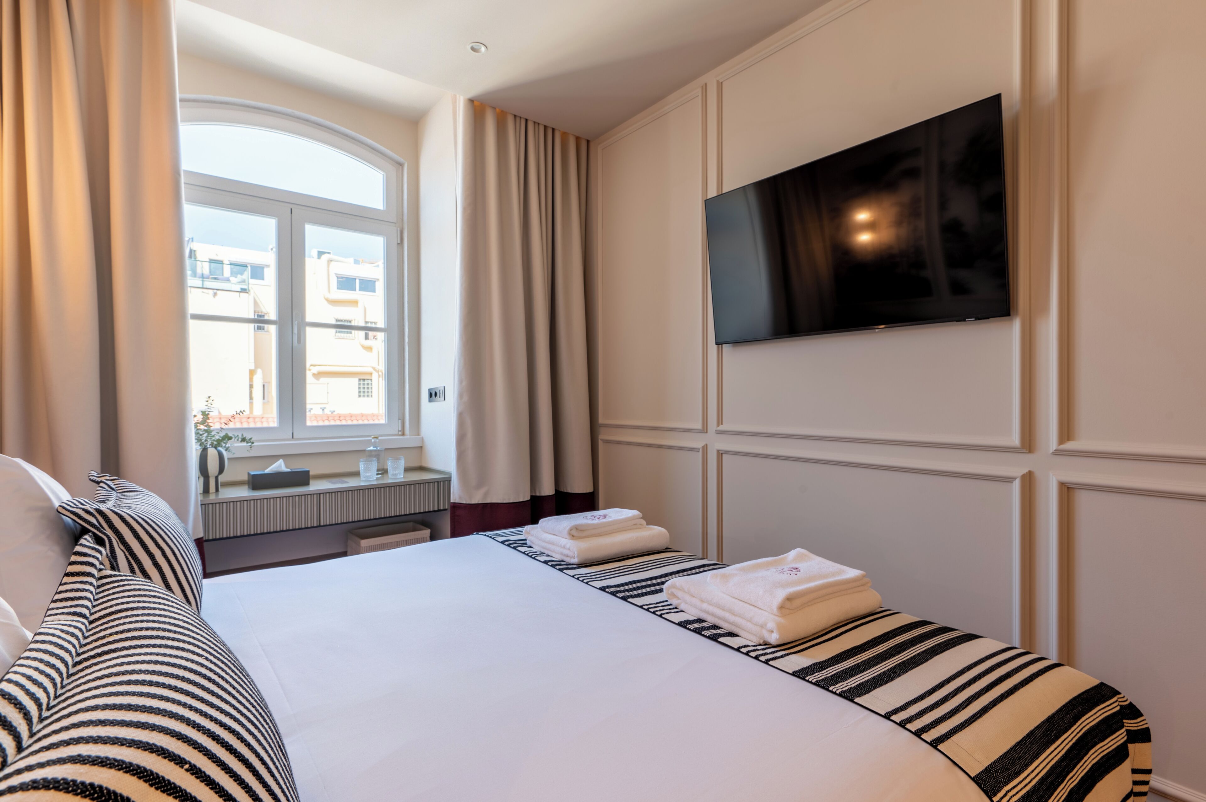 Hotelkamer met een bed, een aan de muur bevestigde televisie, een raam met gordijnen en uitzicht op gebouwen.