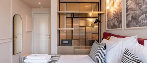Habitación de hotel con cama, mesita de noche, espejo, armario, estante con objetos decorativos y lámpara de pared.