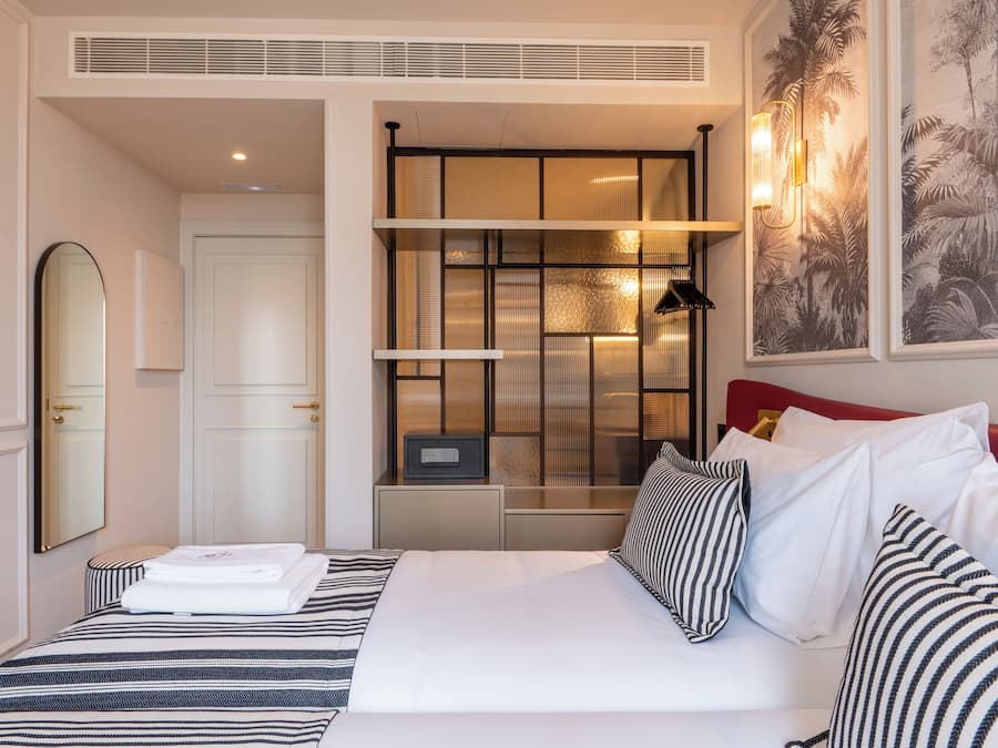 Quarto de hotel com cama, mesa de cabeceira, espelho, guarda-roupa, prateleira com objetos decorativos e uma luminária na parede.