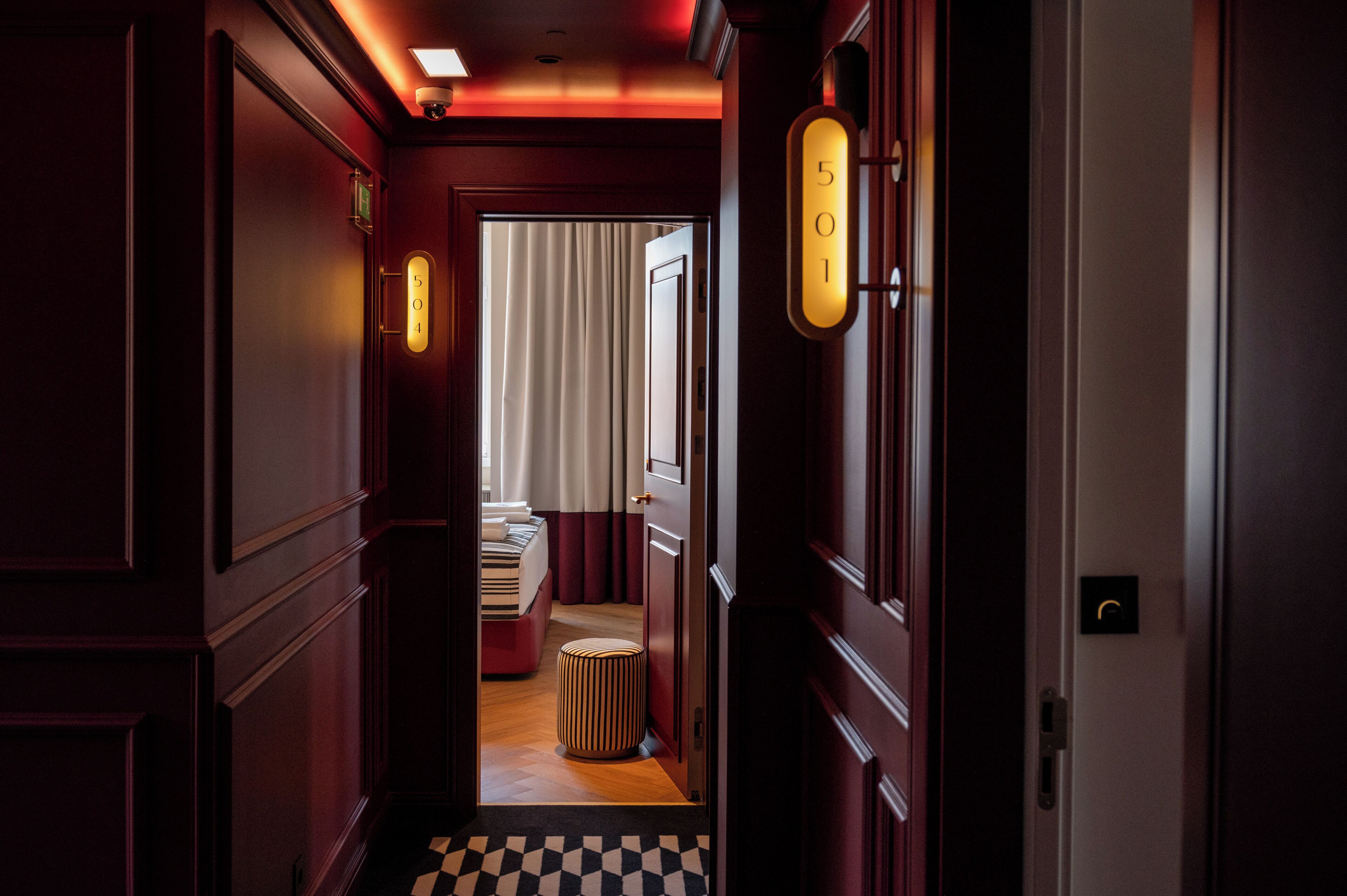 A hotel corridor with a checkered floor, a bed with striped bedding, and a red ottoman.