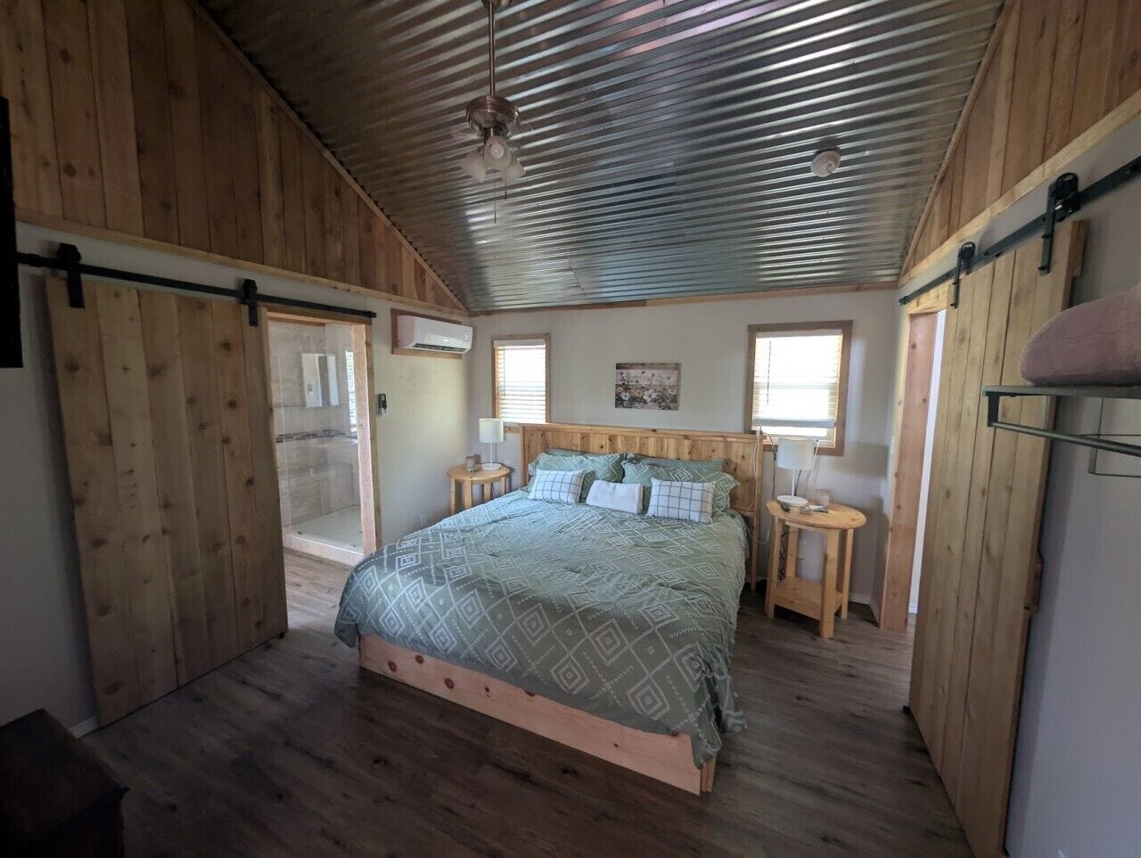 A bedroom with a wooden floor, a bed with a patterned bedspread, two bedside tables with lamps, and a ceiling fan.