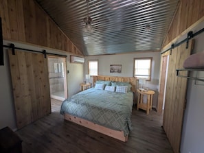 Un dormitorio con suelo de madera, una cama con colcha estampada, dos mesitas de noche con lámparas y un ventilador de techo.