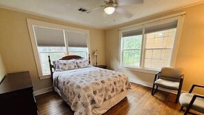 A bedroom with a bed, two windows, a ceiling fan, and a chair. - The Alumni House at Harding (Searcy)