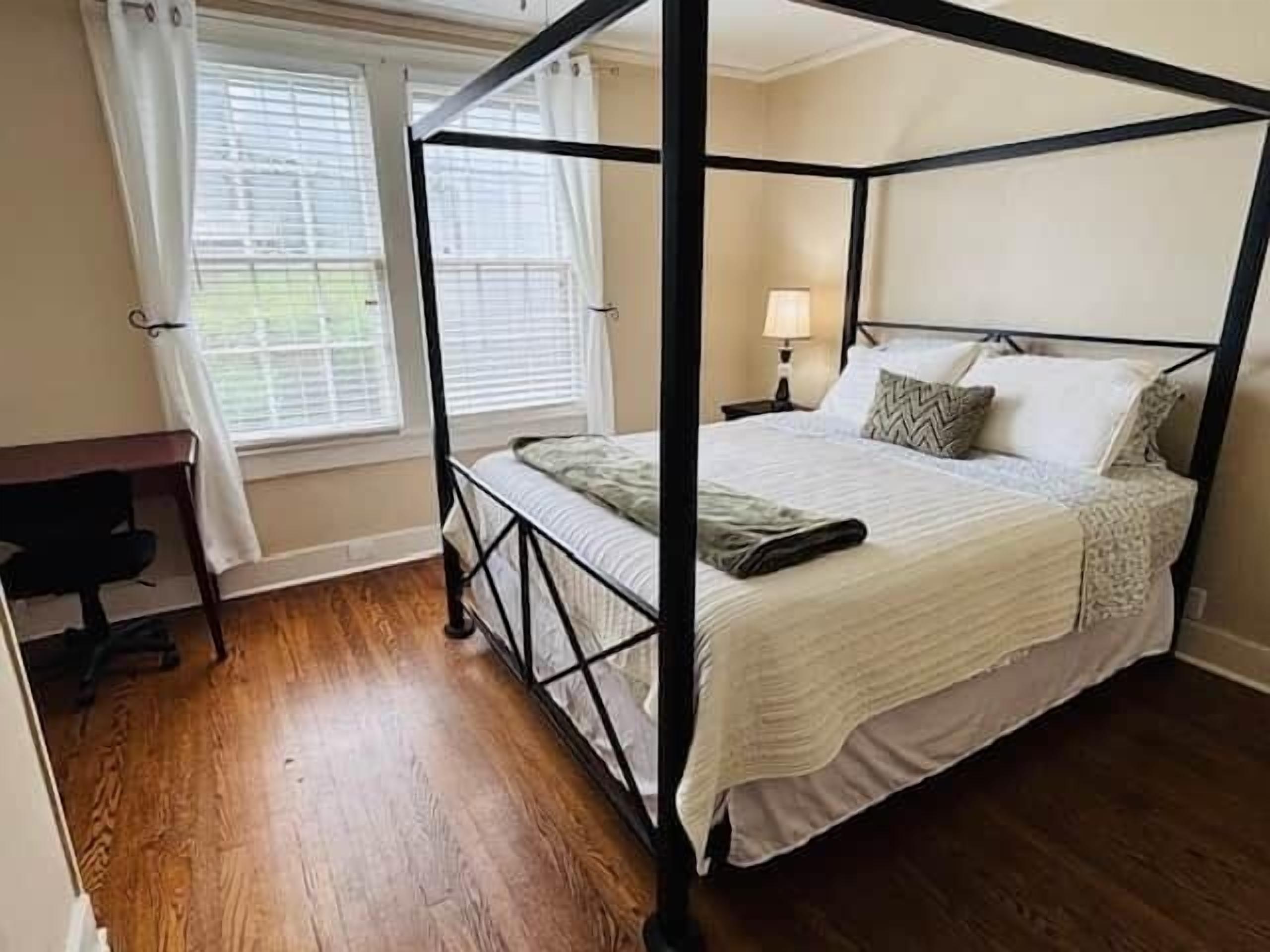 A four-poster bed with a white bedspread and pillows, a desk with a chair, and a window with curtains.