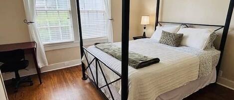 A four-poster bed with a white bedspread and pillows, a desk with a chair, and a window with curtains.