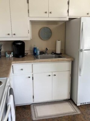 A compact kitchen with white cabinets, a sink, a coffee maker, and a refrigerator. - The Alumni House at Harding (Searcy)