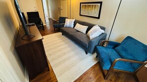 A living room with a sofa, a blue chair, a wooden coffee table, and a standing lamp. - The Alumni House at Harding (Searcy)
