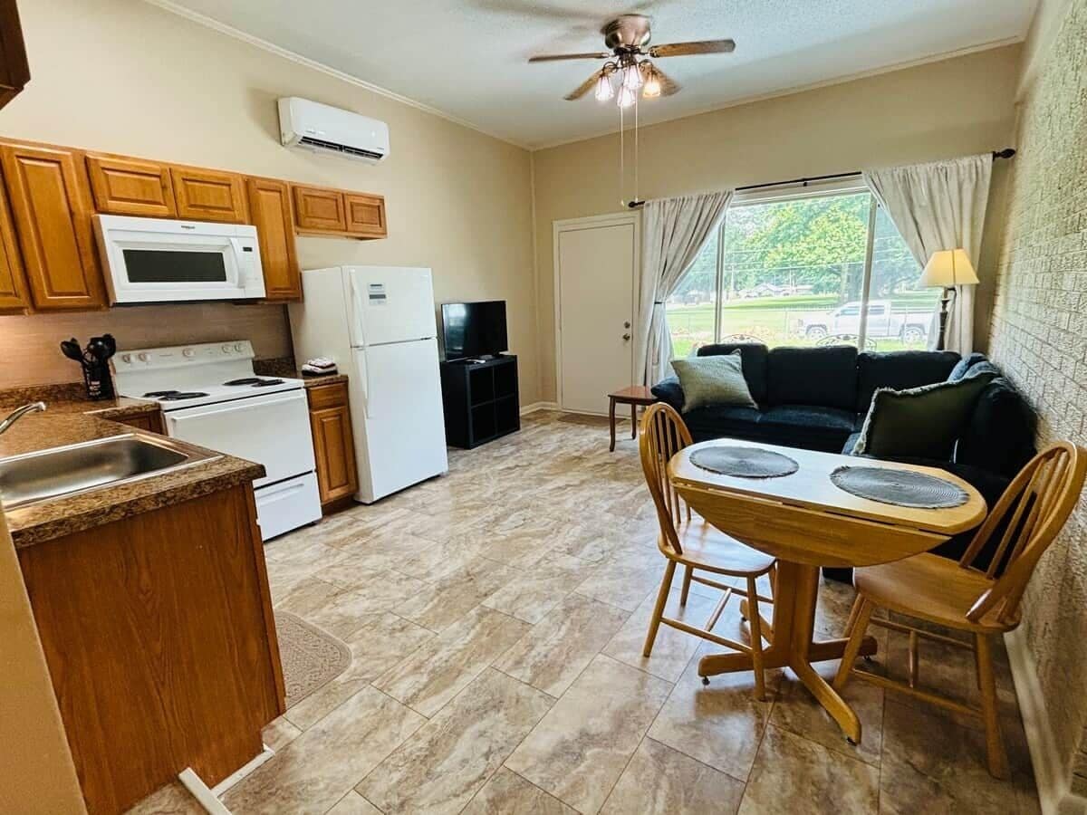 Cocina compacta con microondas, estufa y refrigerador. Hay una zona de comedor con mesa y sillas, y una ventana con cortinas.