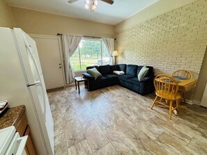 A living room with a sectional sofa, a dining table with chairs, a refrigerator, and a window with curtains.
