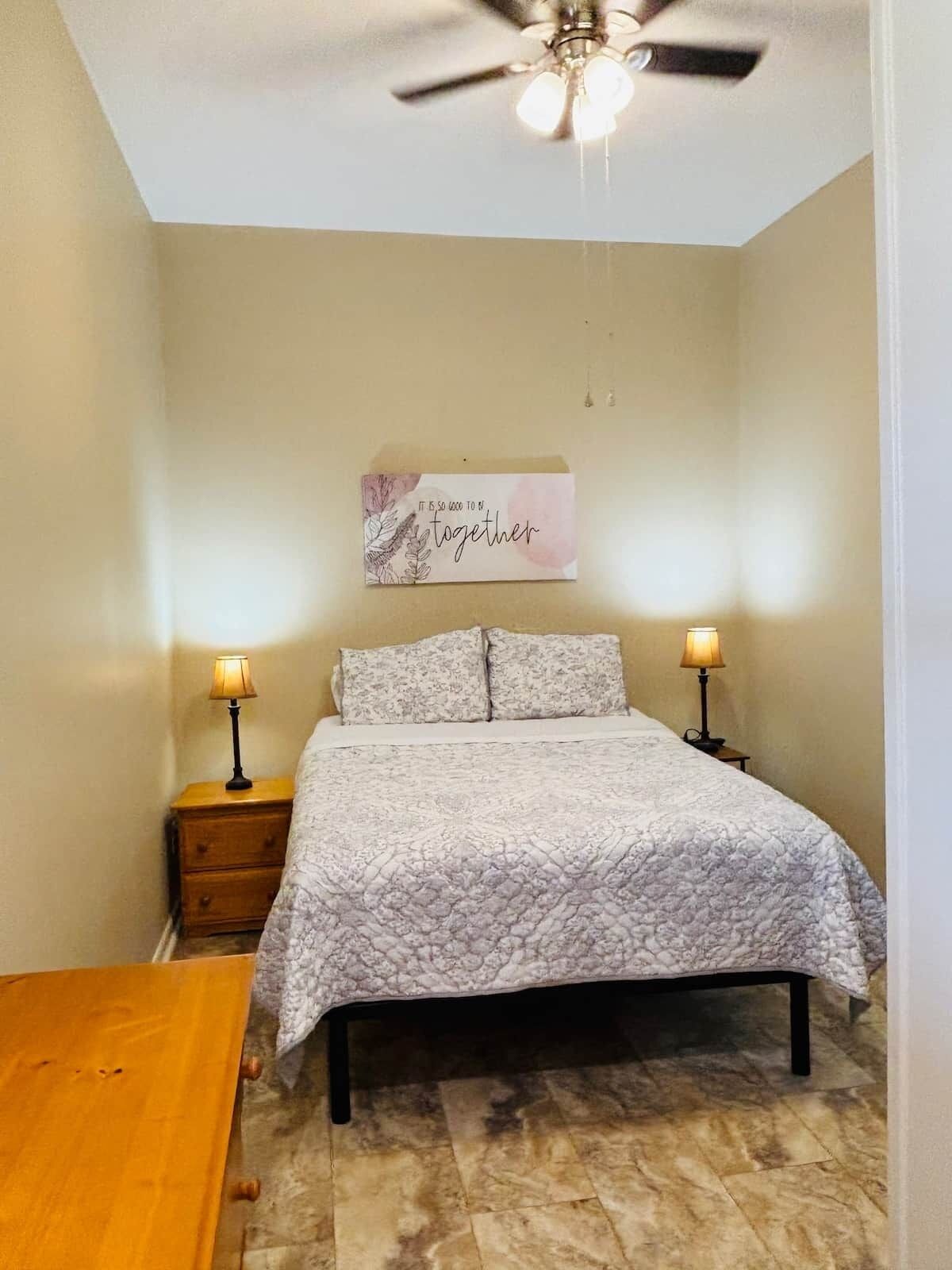 Un dormitorio con cama, dos lámparas de noche, un ventilador de techo, una mesita de noche y una decoración en la pared.
