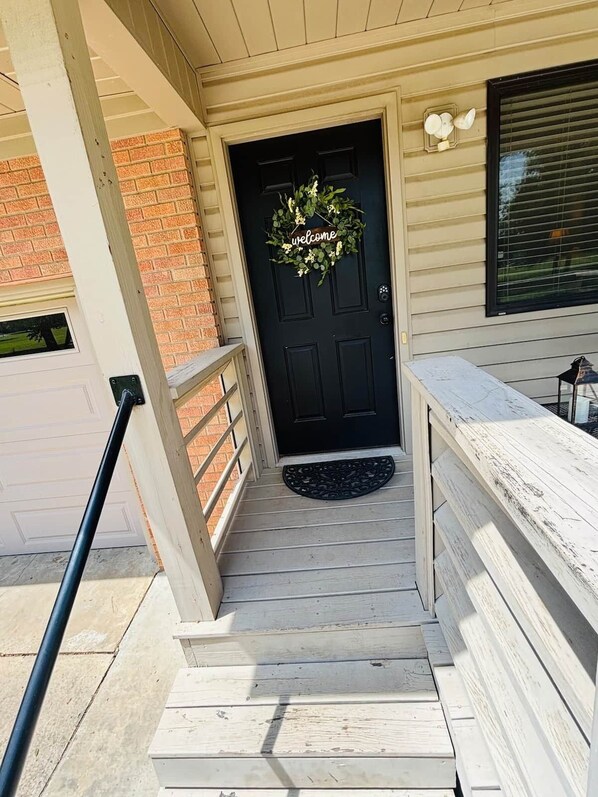 A front porch with a black door, a welcome wreath, and a bench.