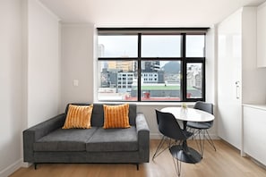 A modern living room with a grey sofa, two orange cushions, a round dining table with black chairs, and a large window with a city view. - Stylish | Roof Top Pool | Studio Apartment | The Tokyo (Cape Town)