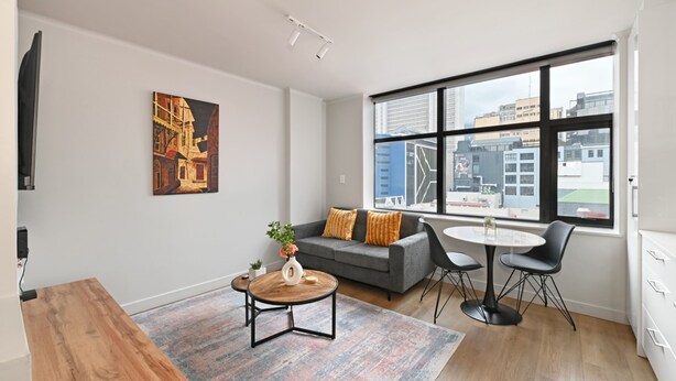 A modern living room with a grey sofa, a coffee table, and a dining area with chairs. There is a large window with a city view. - Stylish | Roof Top Pool | Studio Apartment | The Tokyo (Cape Town)