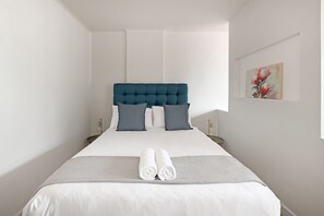 Ein Hotelzimmer mit einem großen Bett, zwei Nachttischen, einem an der Wand hängenden Bild und einem ordentlich bezogenen Bett mit zwei zusammengerollten Handtüchern.