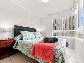 Quarto com cama, mesa de cabeceira, luminária, janela com vista para edifícios altos e manta vermelha na cama.