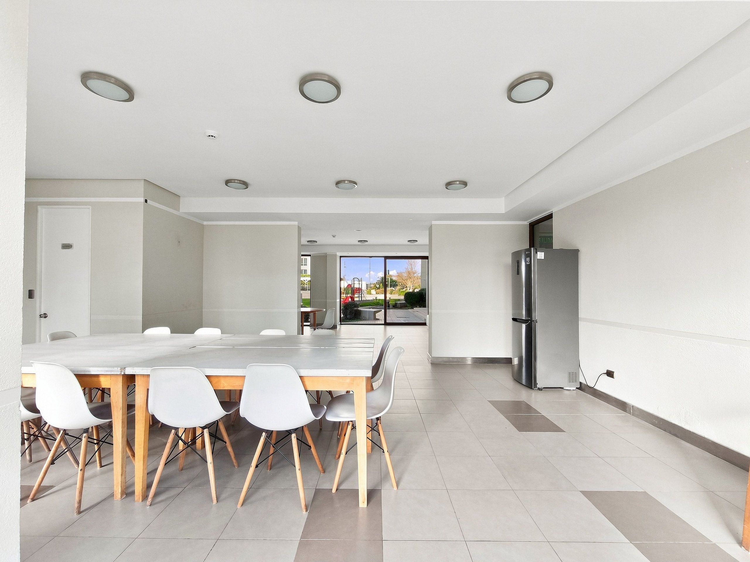 A modern dining area with a long table and chairs, a refrigerator, and a view through a glass door to an outdoor area.