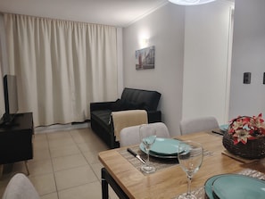 Ein Wohnzimmer mit einer Couch, einem Esstisch gedeckt mit Tellern und Gläsern, einem Fernseher auf einem Lowboard und einem Gemälde an der Wand.