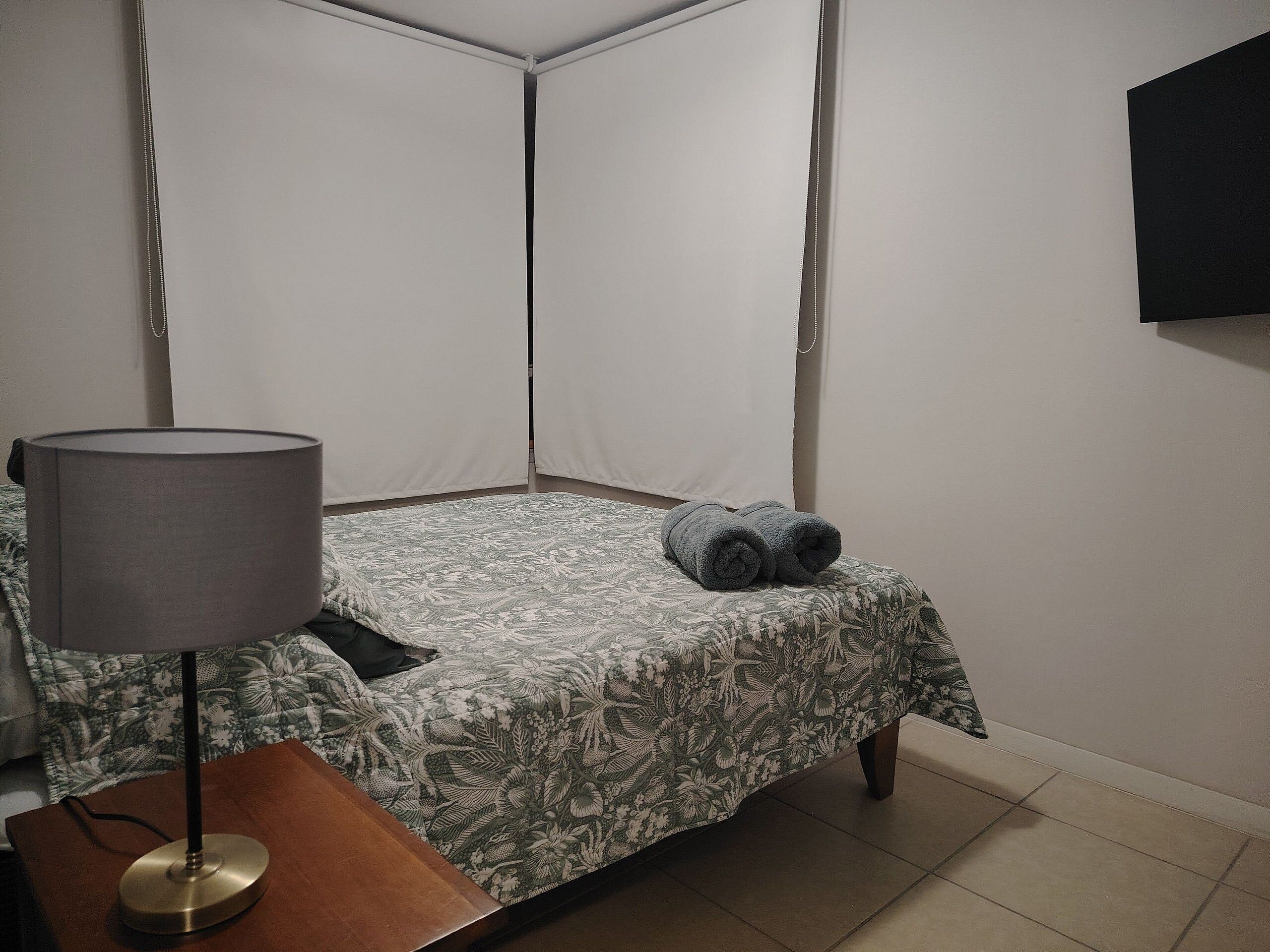 A small, neatly made room with a single bed, a bedside table with a lamp, and a wall-mounted TV.