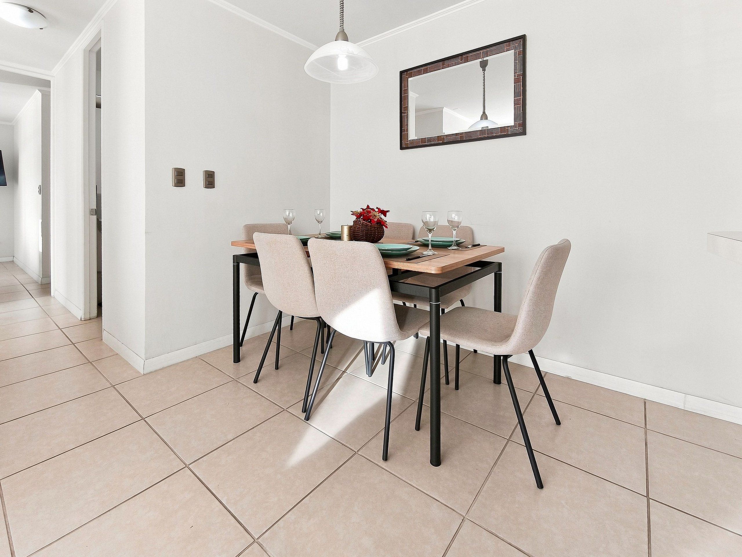 A dining area with a table set for two, chairs, a hanging light fixture, and a wall-mounted mirror.