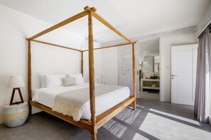 Una cama con dosel, cubierta con una colcha y almohadas blancas, una lámpara de noche, un otoman de almacenamiento redondo y un baño con lavabo y espejo.