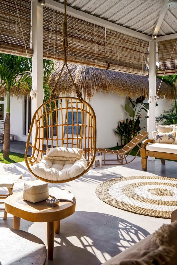 Un espacio al aire libre tropical con una silla colgante, mesas redondas y sombrillas de paja.