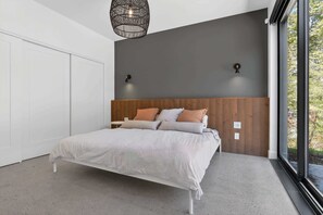 Quarto moderno com uma cama grande, cabeceira de madeira, duas luzes fixadas na parede e vista para o verde através de uma janela.