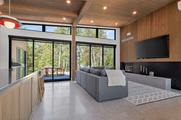 A modern interior with a grey sofa, a flat-screen TV, and large windows overlooking a forest.
