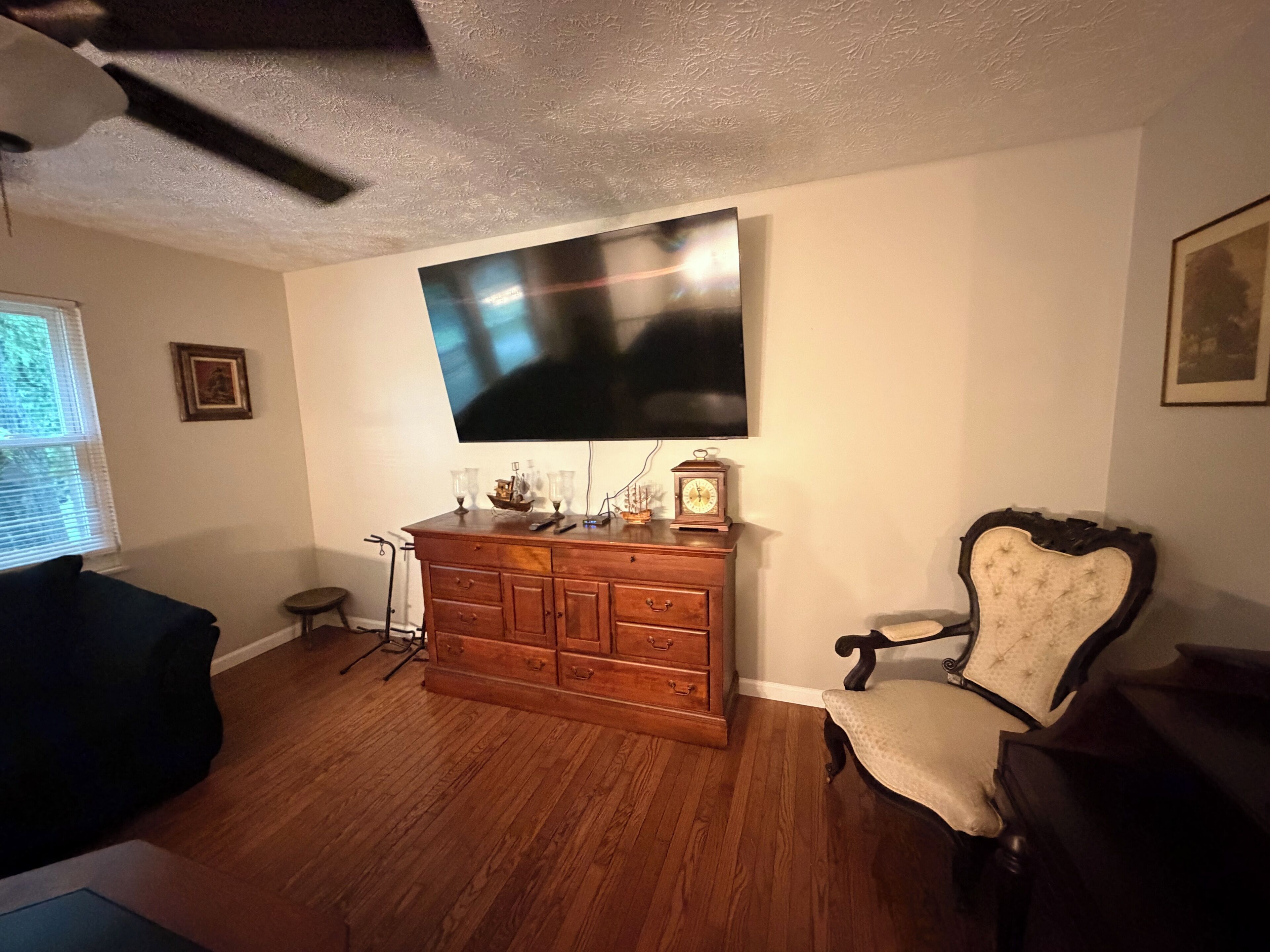 Una sala de estar con un mueble de madera, un televisor montado en la pared, una silla y un ventilador de techo.