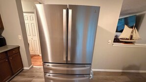 A stainless steel refrigerator in a kitchen with wooden flooring and a white door in the background.