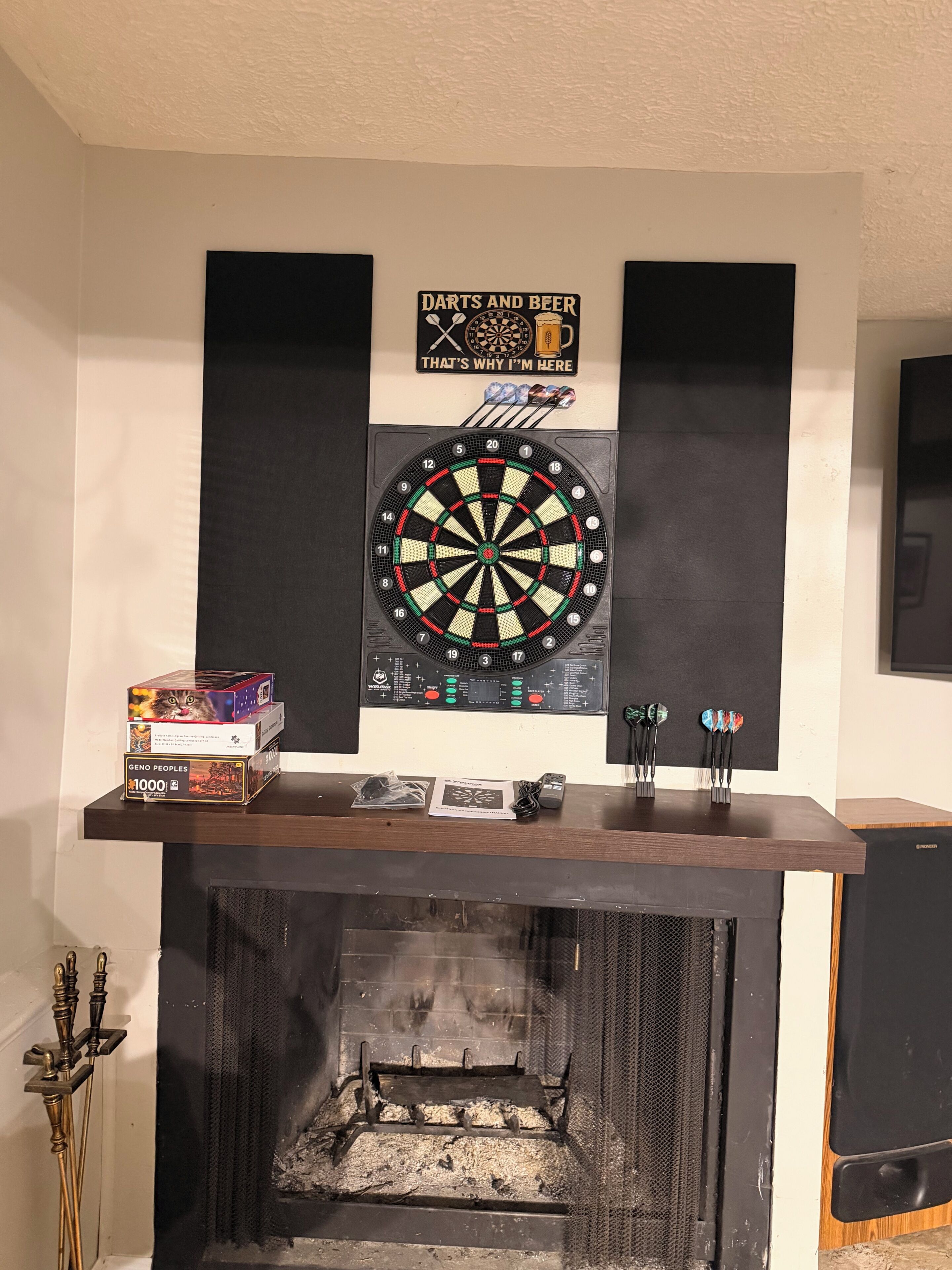 A dartboard mounted on a wall with a sign above it, a fireplace with a mantel, and a speaker to the right.