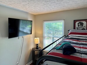 Un dormitorio con cama, televisor montado en la pared, ventana con persianas y lámpara en una mesita de noche.
