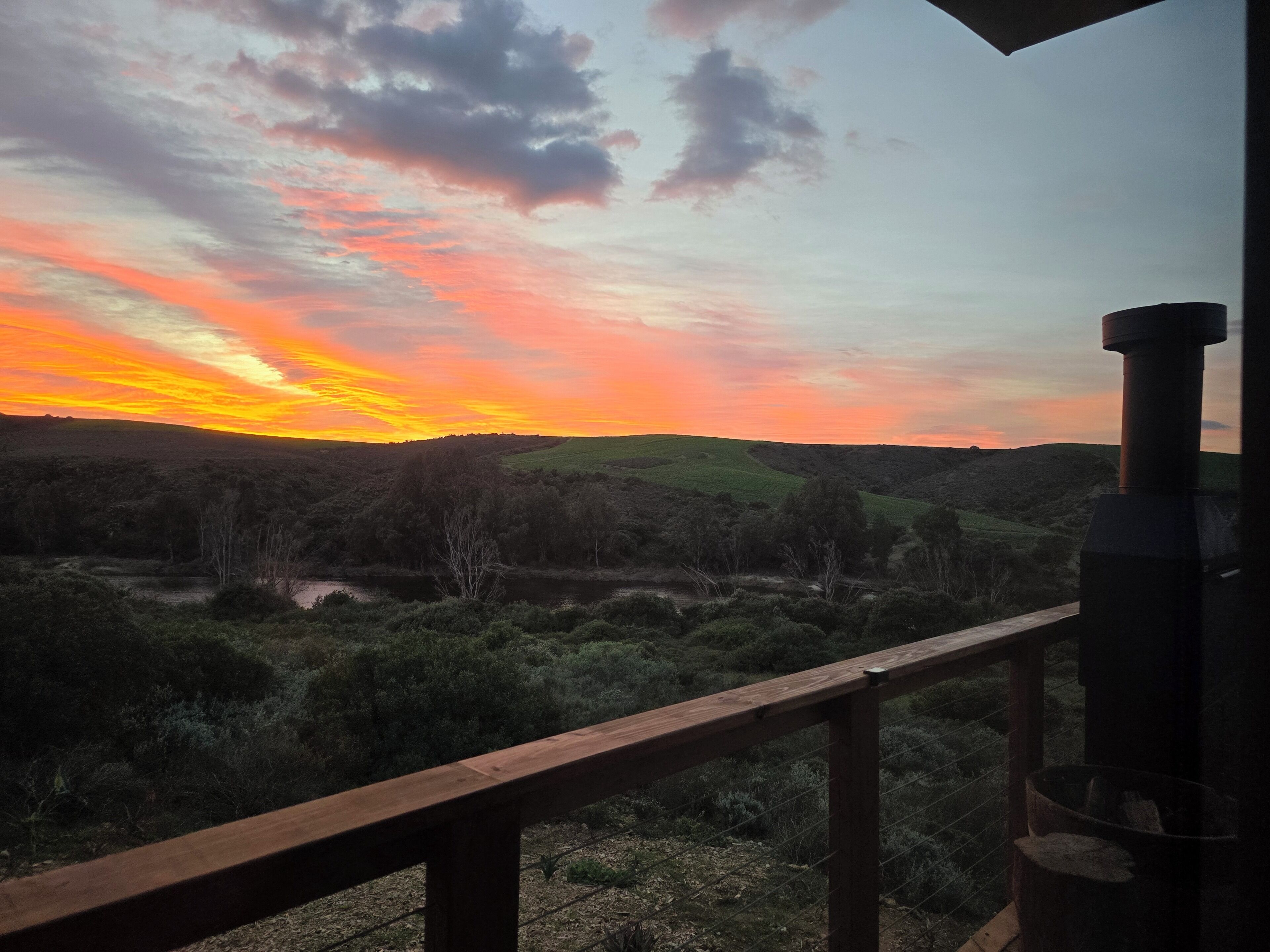 A sunset view from a balcony with a wooden railing, overlooking a hilly landscape with sparse vegetation.