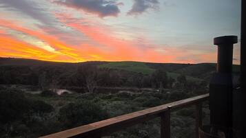 A sunset view from a balcony with a wooden railing, overlooking a hilly landscape with sparse vegetation.
