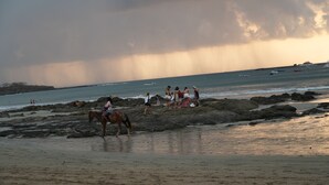 A beach with people and horses, rocky terrain, and boats in the water.