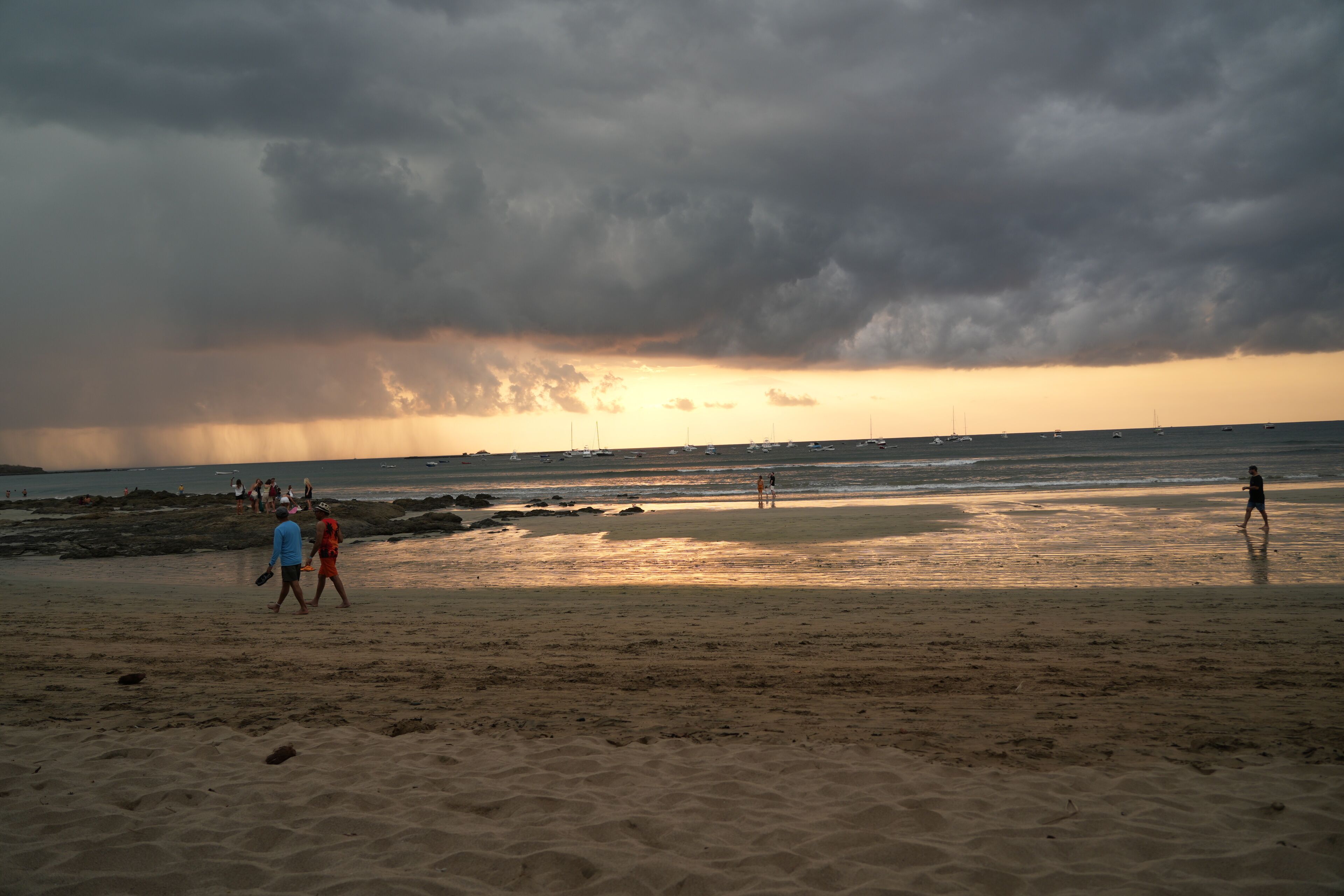 A beach with people walking, boats in the water, and a cloudy sky.
